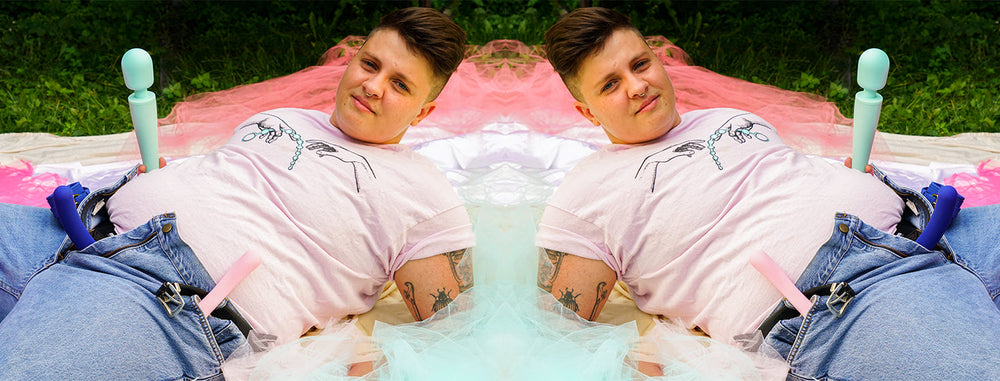 Photo of June Pilote lying down, holding wand vibrator and wearing strap on with dildo attached